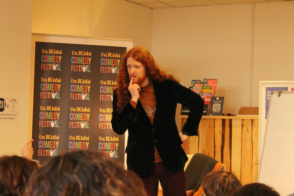 Comedian Alasdair Beckett King, a tall man with long red hair, stands quizzically at the front of a bookshop with a The UK Kids' Comedy Festival banner as a backdrop. At the bottom of the image you can see the backs of children's heads looking at the comedian.