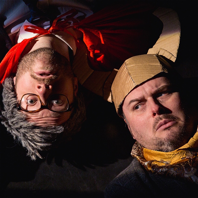 The face of a young(ish) man in a grey wig, pretending to be old, hangs upside down in a red cape. Next to his face is an upright man's face. He wears a cravat, sporting genuinely grey stubble and has a viking helmet on his head made of cardboard.